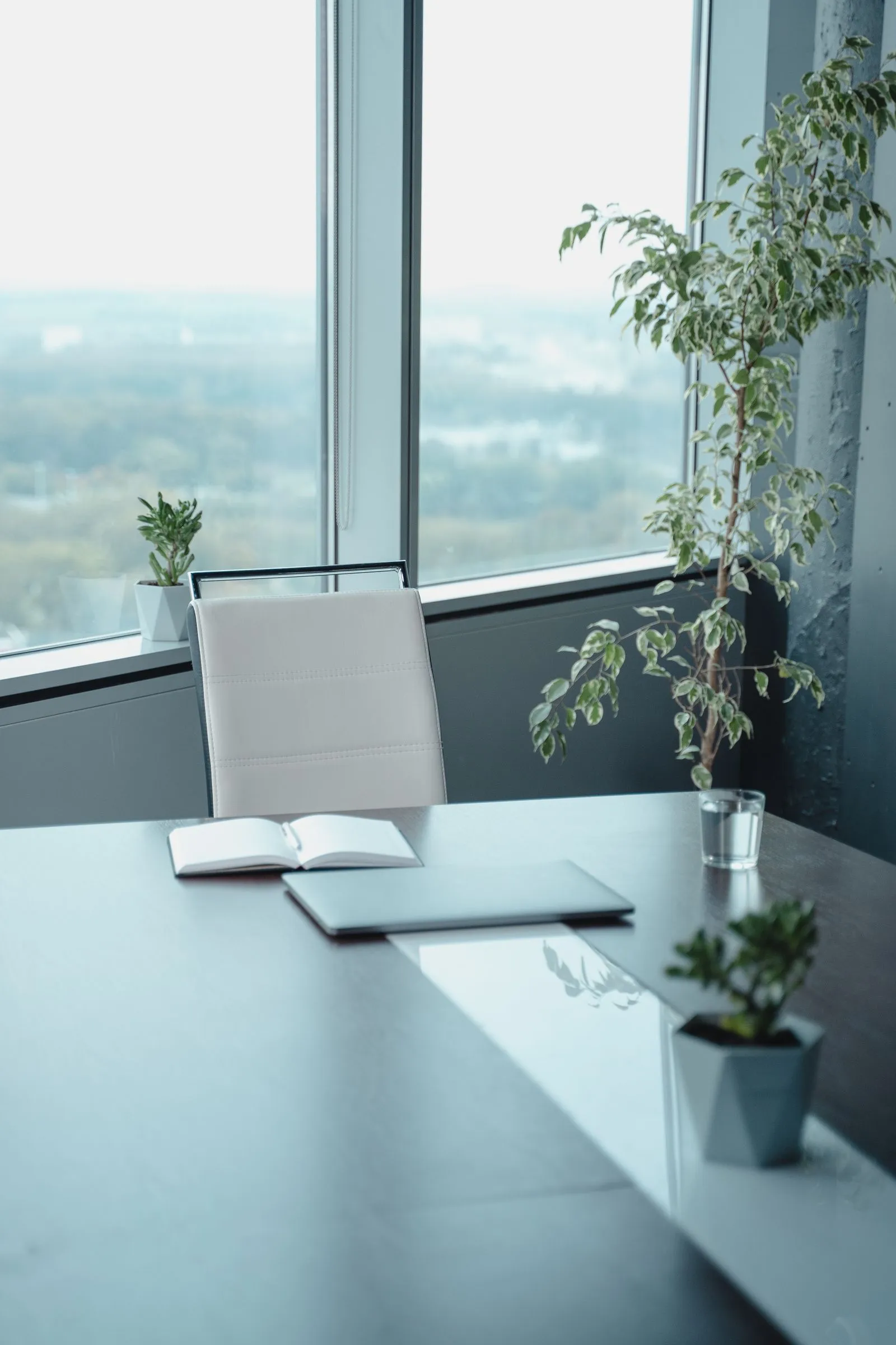 Mesa de escritório com caderno aberto e notebook diante de uma janela ampla